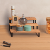 Dreistufiges Holzregal mit schwarzen Metallfüßen auf hellem Holztisch, darauf eine runde Glasflasche, eine rechteckige Flasche, eine hohe Sprühdose und zwei grüne Tiegel; rechts ein Glasbehälter mit orangefarbenen Kapseln; vorne links zwei schlanke S