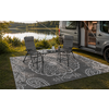 Fourgon aménagé gris avec porte latérale ouverte et auvent déployé ; devant un tapis d'extérieur à motif, deux chaises de camping pliantes et une petite table avec une carafe et des verres, au bord d'un lac avec des arbres en arrière-plan