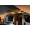 Camping-car argenté au coucher du soleil, auvent déployé avec bande lumineuse, porte coulissante ouverte montrant l'intérieur avec banquette et coin cuisine, arbres et montagnes en arrière-plan