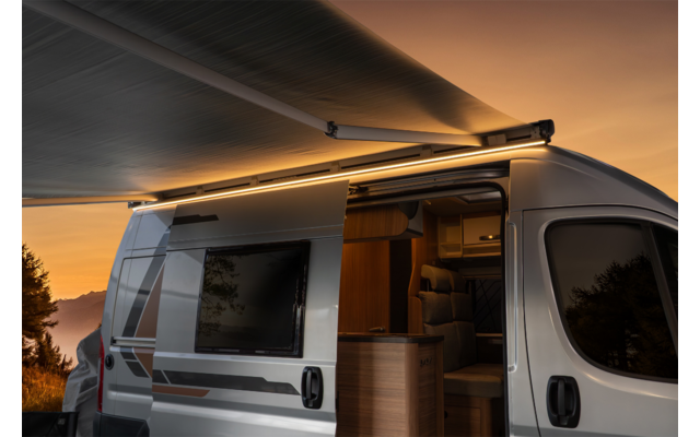 Camping-car argenté au coucher du soleil, auvent déployé avec bande lumineuse, porte coulissante ouverte montrant l'intérieur avec banquette et coin cuisine, arbres et montagnes en arrière-plan