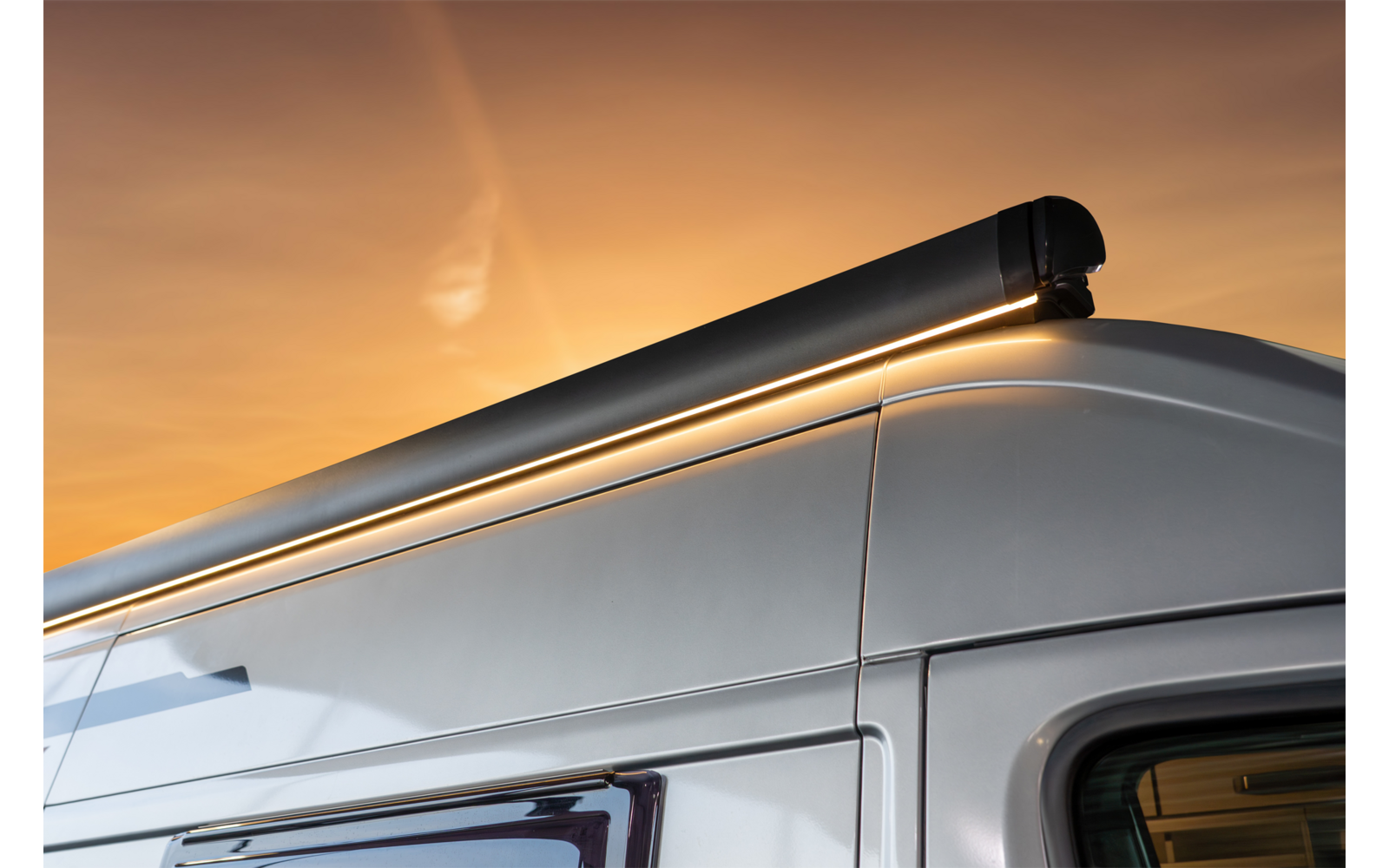 Partie supérieure d'un camping-car blanc avec un store fermé fixé au bord du toit, ciel orange en arrière-plan