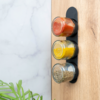 Drie kleine glazen potjes met rode, gele en groene inhoud op een zwarte houder aan een houten plaat; links een marmeren wand en groene plantbladeren