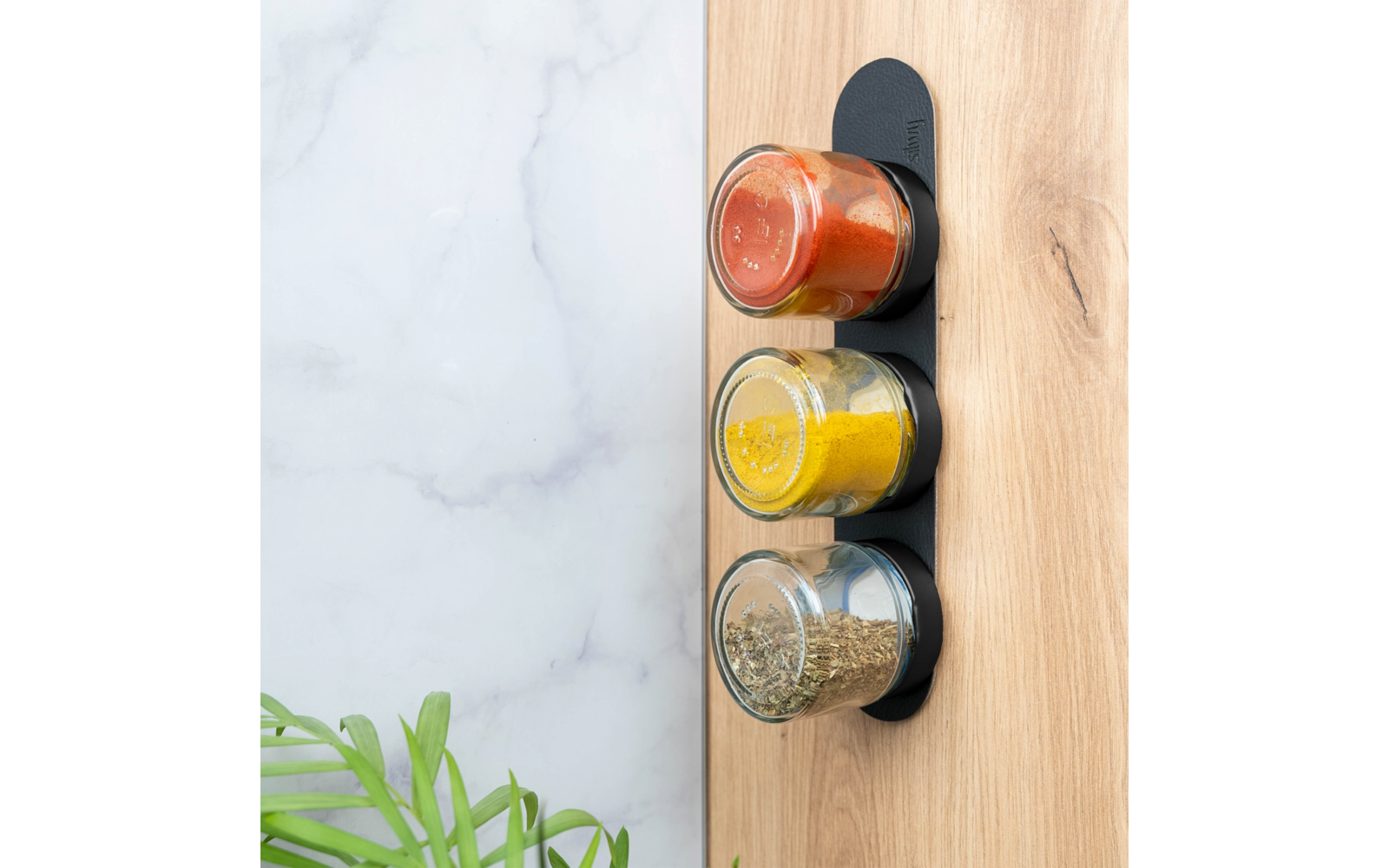 Drie kleine glazen potjes met rode, gele en groene inhoud op een zwarte houder aan een houten plaat; links een marmeren wand en groene plantbladeren