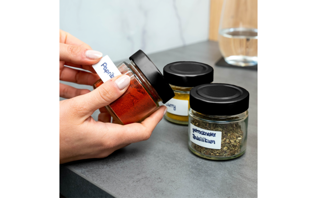 Twee handen houden een klein glazen potje met rood poeder en een wit etiket met de tekst "Paprika"; op het aanrecht twee glazen potjes met zwarte deksels, één met geel poeder, één met gedroogde groene kruiden en een etiket met de tekst "getrockneter Basilikum"; op de achtergrond een glas water