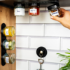 Hand pakt een klein glazen potje met schroefdeksel; meerdere gelabelde glazen potjes onder een plank en op een verticale magnetische strook; witte tegels, houten paneel links, zwarte haak met sleutelhanger, plantenbladeren rechtsonder