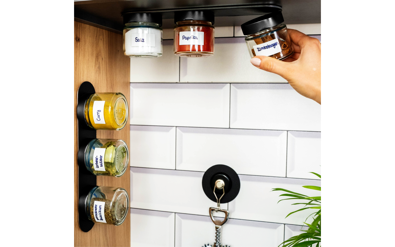 Hand pakt een klein glazen potje met schroefdeksel; meerdere gelabelde glazen potjes onder een plank en op een verticale magnetische strook; witte tegels, houten paneel links, zwarte haak met sleutelhanger, plantenbladeren rechtsonder