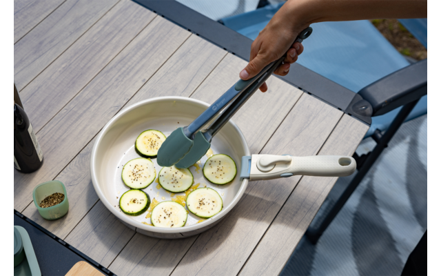 Mano con pinzas de cocina sostiene rodajas de calabacín en una sartén blanca sobre una mesa de madera; frasco de especias, botella y silla con cojín azul visibles