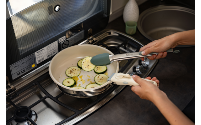 Dos manos sujetan unas pinzas de cocina sobre una sartén con rodajas de calabacín en aceite en una cocina de gas; fregadero y encimera visibles al fondo