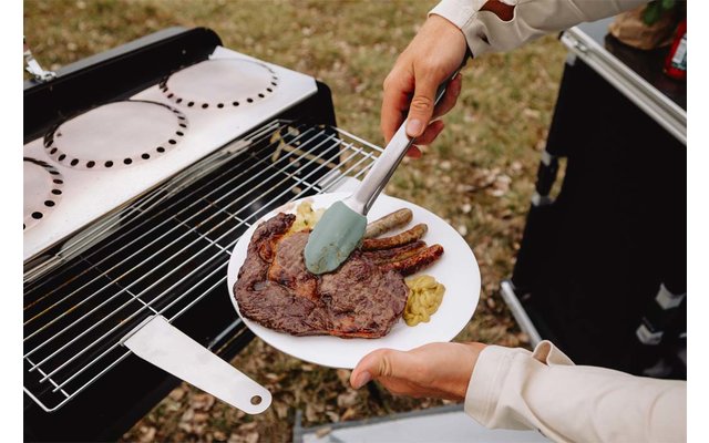 Teller mit gegrilltem Steak, mehreren Würstchen und einem Klecks Senf; eine Hand hält den Teller, eine andere legt mit einer Spachtel Fleisch vom Grill; Rasen im Hintergrund
