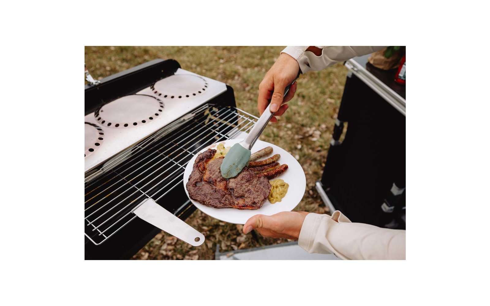 Teller mit gegrilltem Steak, mehreren Würstchen und einem Klecks Senf; eine Hand hält den Teller, eine andere legt mit einer Spachtel Fleisch vom Grill; Rasen im Hintergrund