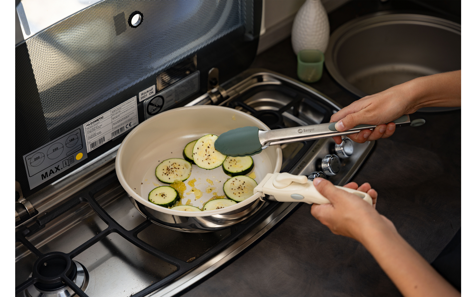Zwei Hände halten eine Küchenzange über einer Pfanne mit Zucchinischeiben in Öl auf einem Gasherd; Spüle und Arbeitsfläche im Hintergrund