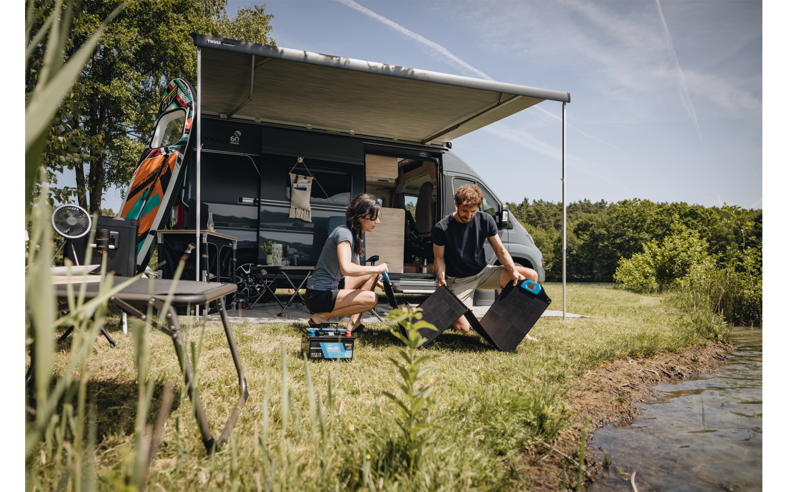 Autocaravana con toldo desplegado en la orilla de un lago. Dos personas arrodilladas en la hierba delante del vehículo junto a una caja abierta y equipo de camping; dos tablas largas en el techo, sillas y una mesa pequeña al lado; orilla y árboles al fondo