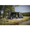 Autocaravana gris con toldo desplegado en la orilla de un lago; dos personas arrodilladas sobre una esterilla plegable gris junto a varios objetos pequeños; una tabla naranja apoyada en el vehículo; hierba, juncos, árboles y cielo azul con estelas de condensación