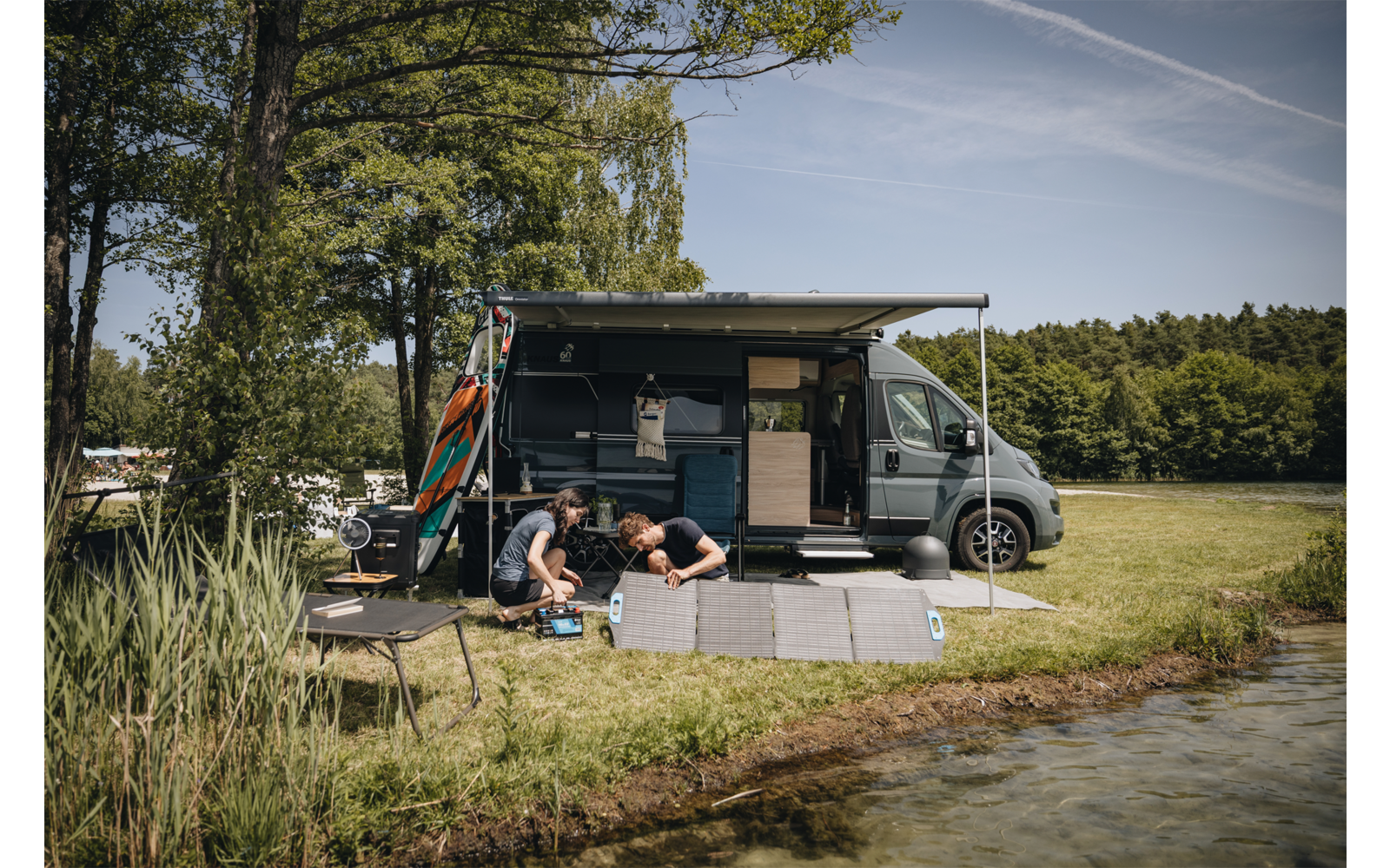 Autocaravana gris con toldo desplegado en la orilla de un lago; dos personas arrodilladas sobre una esterilla plegable gris junto a varios objetos pequeños; una tabla naranja apoyada en el vehículo; hierba, juncos, árboles y cielo azul con estelas de condensación