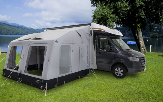 Autocaravana gris con toldo/tienda blanca acoplada sobre césped verde junto a un lago, árbol a la derecha, montañas y cielo nublado al fondo