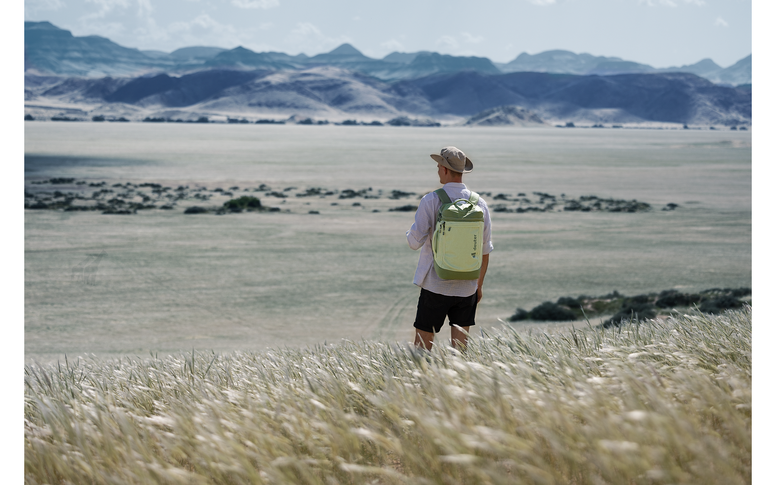 Person mit hellgrünem Rucksack und Kappe, mit dem Rücken zur Kamera, steht im hohen Gras vor einer weiten Ebene und fernen Bergen