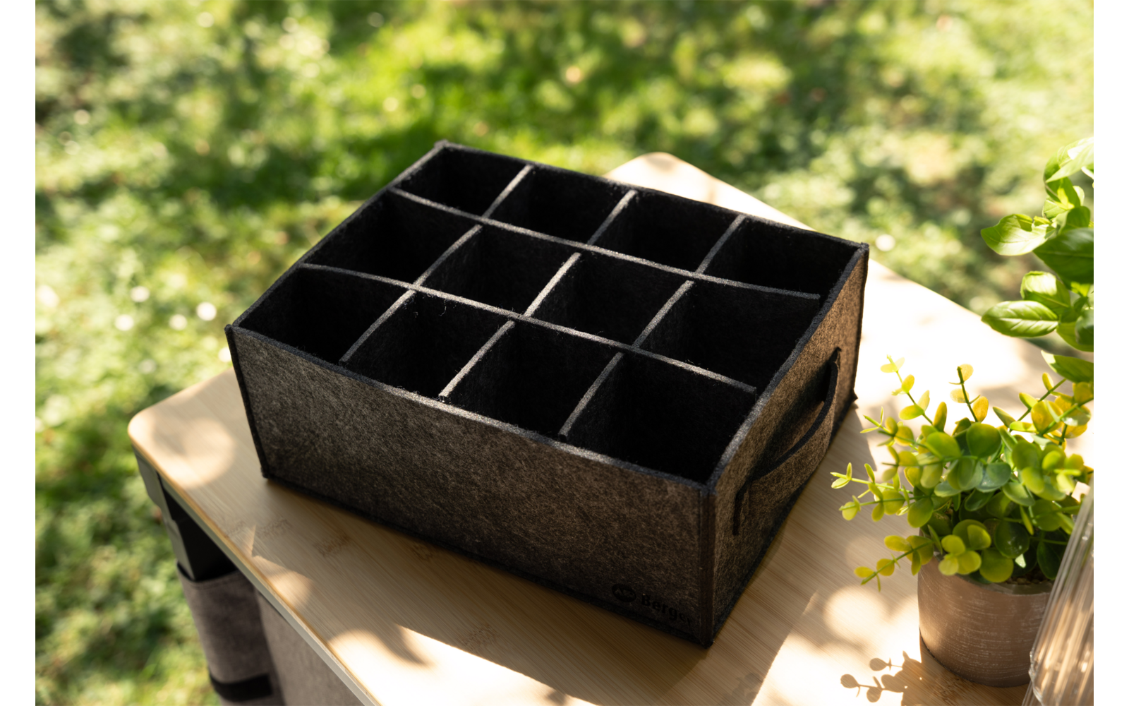 Dunkelgraue Filzbox mit zwölf rechteckigen Fächern auf einem hellen Holztisch im Freien; rechts ein kleiner Pflanzenkübel; unscharfer Rasen im Hintergrund; Sonnenlicht und Schatten