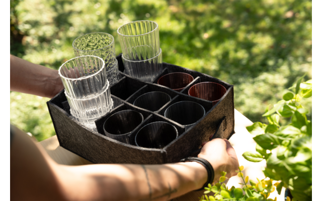 Zwei Unterarme halten ein schwarzes, in neun Fächer unterteiltes Tablett mit drei transparenten, gerillten Gläsern und mehreren dunklen Bechern; verschwommener grüner Gartenhintergrund