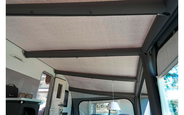 Innenraum mit hellgrauem Stoffdach und drei dunklen Querstreben, rechts Fenster mit halb heruntergelassenem grauen Rollo, links Arbeitsfläche mit schwarzer Tasche, im Hintergrund weiße Wohnmobiltür und hängende weiße Lampe