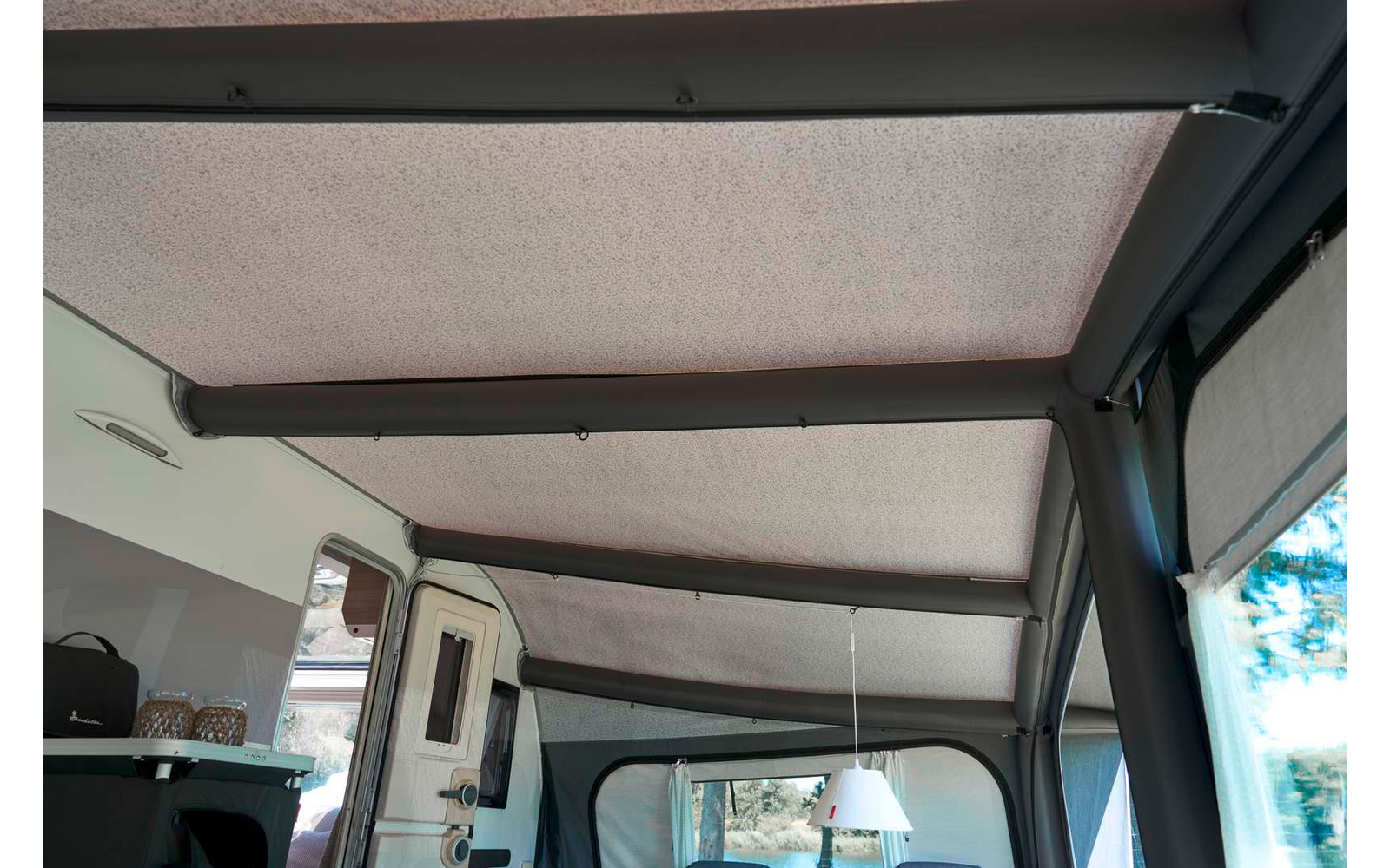 Innenraum mit hellgrauem Stoffdach und drei dunklen Querstreben, rechts Fenster mit halb heruntergelassenem grauen Rollo, links Arbeitsfläche mit schwarzer Tasche, im Hintergrund weiße Wohnmobiltür und hängende weiße Lampe