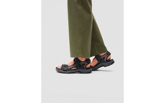 Mollets et pieds portant des sandales de sport noires à brides réglables, pantalons larges couleur olive sur fond gris clair