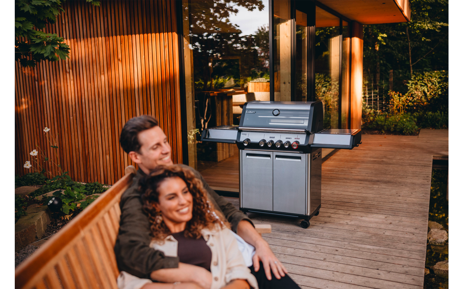 Zwei Personen sitzen auf einer Holzbank auf einer Holzterrasse neben einem Edelstahl-Gasgrill; im Hintergrund Haus mit Holzverkleidung und Pflanzen