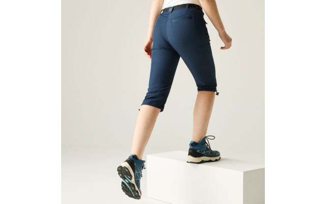 Unterkörper einer Person von der Hüfte abwärts, mit einem Fuß auf einem weißen Quader, dunkelblaue knielange Hose und blaue Schnürschuhe