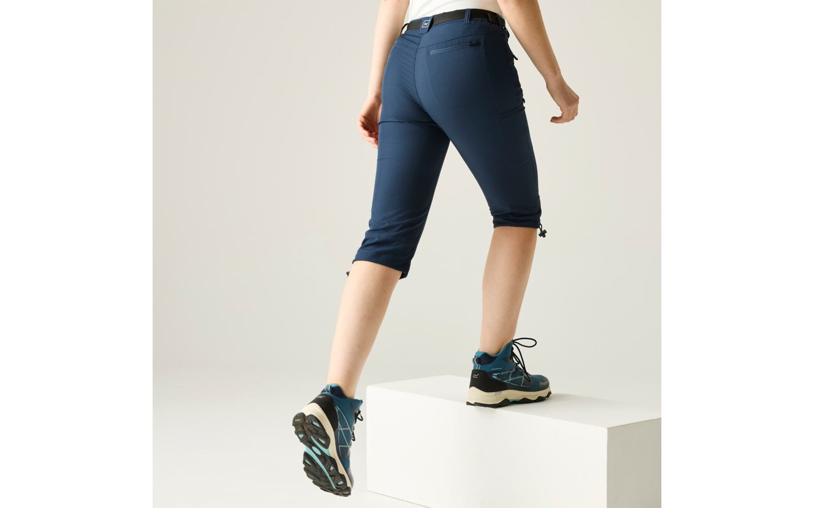 Unterkörper einer Person von der Hüfte abwärts, mit einem Fuß auf einem weißen Quader, dunkelblaue knielange Hose und blaue Schnürschuhe