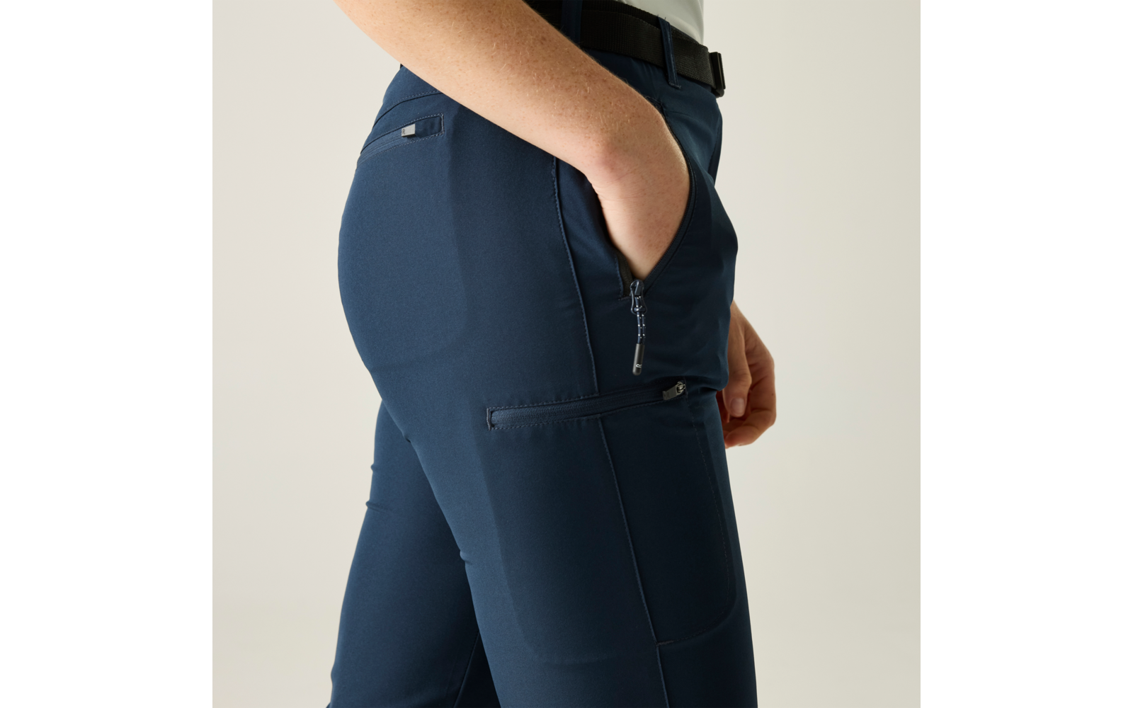Nahaufnahme der Hüfte und Oberschenkel einer Person in dunkelblauen Hosen, rechte Hand in seitlicher Tasche, sichtbarer Reißverschluss und Gürtelschlaufen, heller Hintergrund