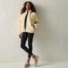 Persona con capelli ricci, giacca in pile beige, leggings neri e scarpe da ginnastica beige, in piedi con le gambe incrociate davanti a una parete chiara