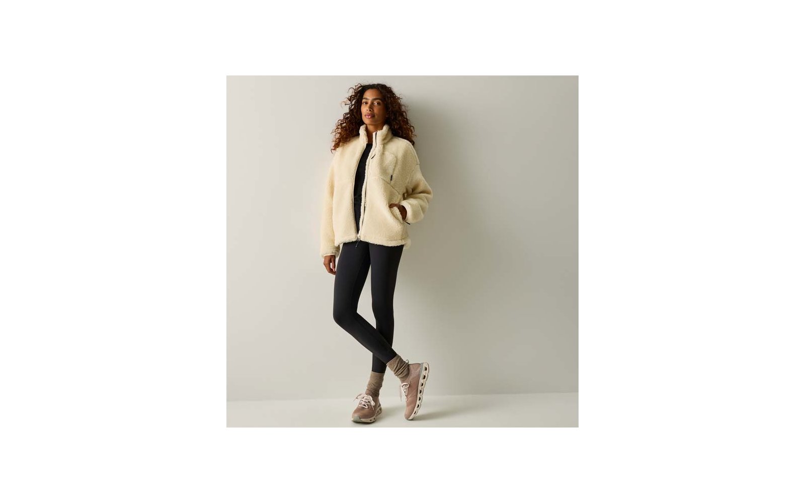 Persona con capelli ricci, giacca in pile beige, leggings neri e scarpe da ginnastica beige, in piedi con le gambe incrociate davanti a una parete chiara