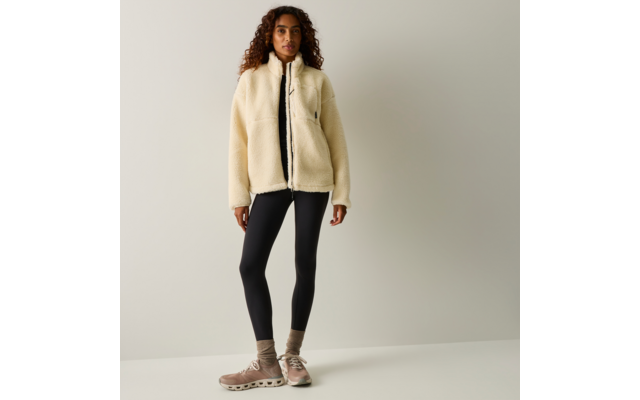 Persona in piedi davanti a uno sfondo neutro che indossa una giacca in pile color crema con cerniera, un top nero, leggings neri, calzini e scarpe da ginnastica beige