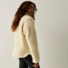 Persona con capelli lunghi e ricci vista da dietro e leggermente di profilo, indossa una giacca in pile crema e leggings neri su sfondo chiaro uniforme
