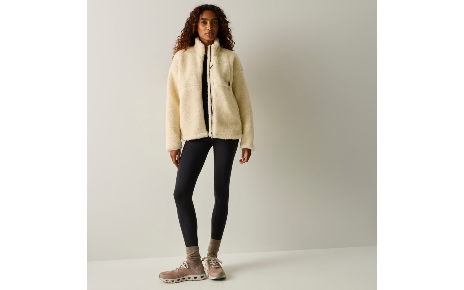 Persona in piedi davanti a uno sfondo neutro che indossa una giacca in pile color crema con cerniera, un top nero, leggings neri, calzini e scarpe da ginnastica beige