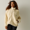 Persona con capelli lunghi e ricci che indossa una giacca in pile beige chiaro con cerniera e due tasche, una mano in una tasca; in piedi davanti a uno sfondo chiaro uniforme
