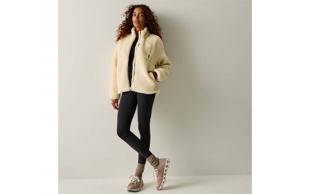 Persona con capelli ricci, giacca in pile beige, leggings neri e scarpe da ginnastica beige, in piedi con le gambe incrociate davanti a una parete chiara