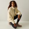 Una persona con capelli ricci accovacciata su un ginocchio, indossa una giacca in pile crema, leggings neri e sneakers beige su sfondo neutro