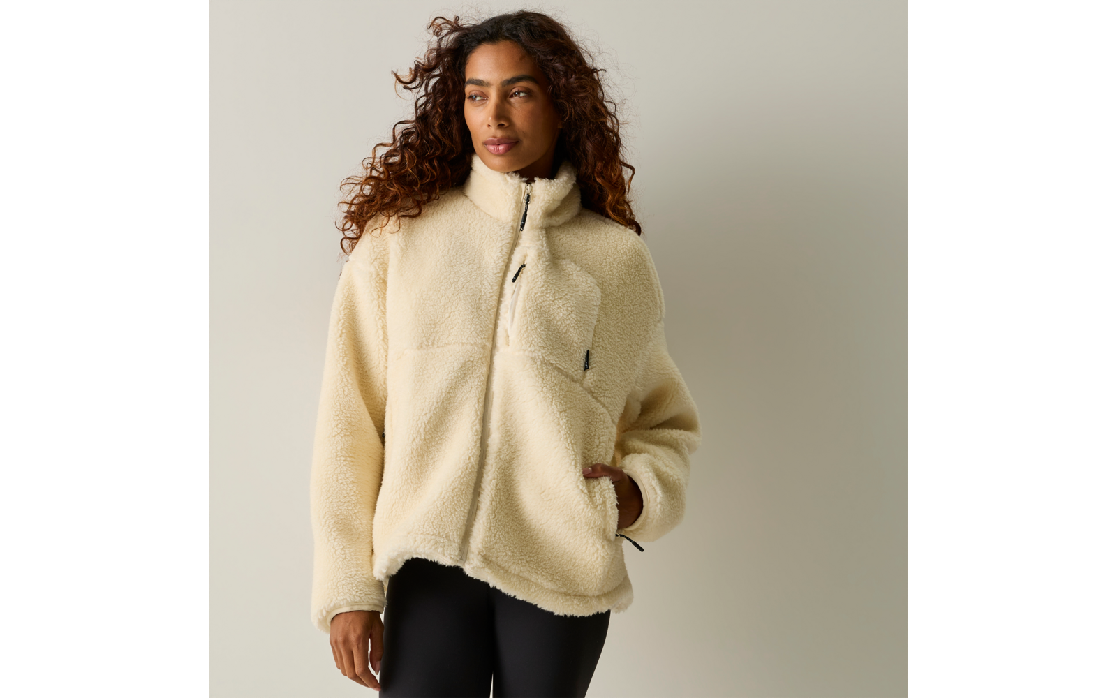 Persona con capelli lunghi e ricci che indossa una giacca in pile beige chiaro con cerniera e due tasche, una mano in una tasca; in piedi davanti a uno sfondo chiaro uniforme