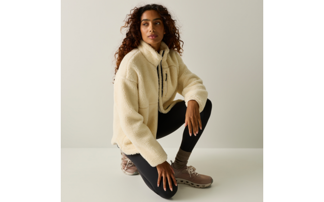 Una persona con capelli ricci accovacciata su un ginocchio, indossa una giacca in pile crema, leggings neri e sneakers beige su sfondo neutro