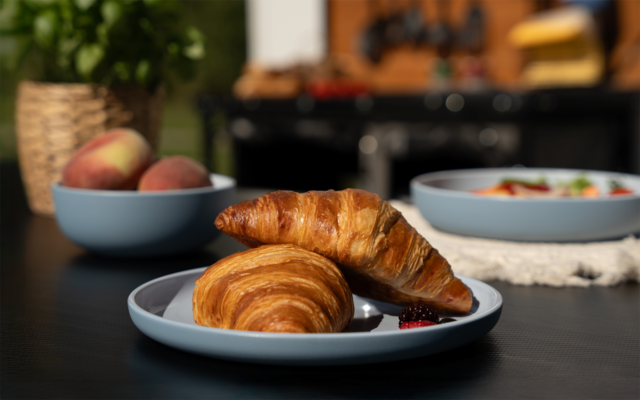 Zwei Croissants auf einem hellblauen Teller auf einem dunklen Tisch; Marmelade neben dem Teller; im Hintergrund zwei blaue Schalen mit Pfirsichen und einem Salat, unscharfe Küchenutensilien