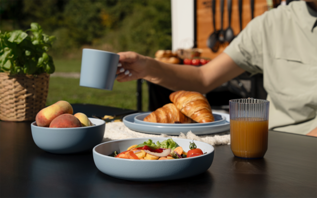 Schwarzer Tisch im Freien mit blauem Geschirr: Schale mit Pfirsichen, Schale mit geschnittenem Gemüse und Tomaten, Teller mit Croissants, Glas Orangensaft, eine Hand hält eine blaue Tasse; Pflanzen und Küchenutensilien im Hintergrund