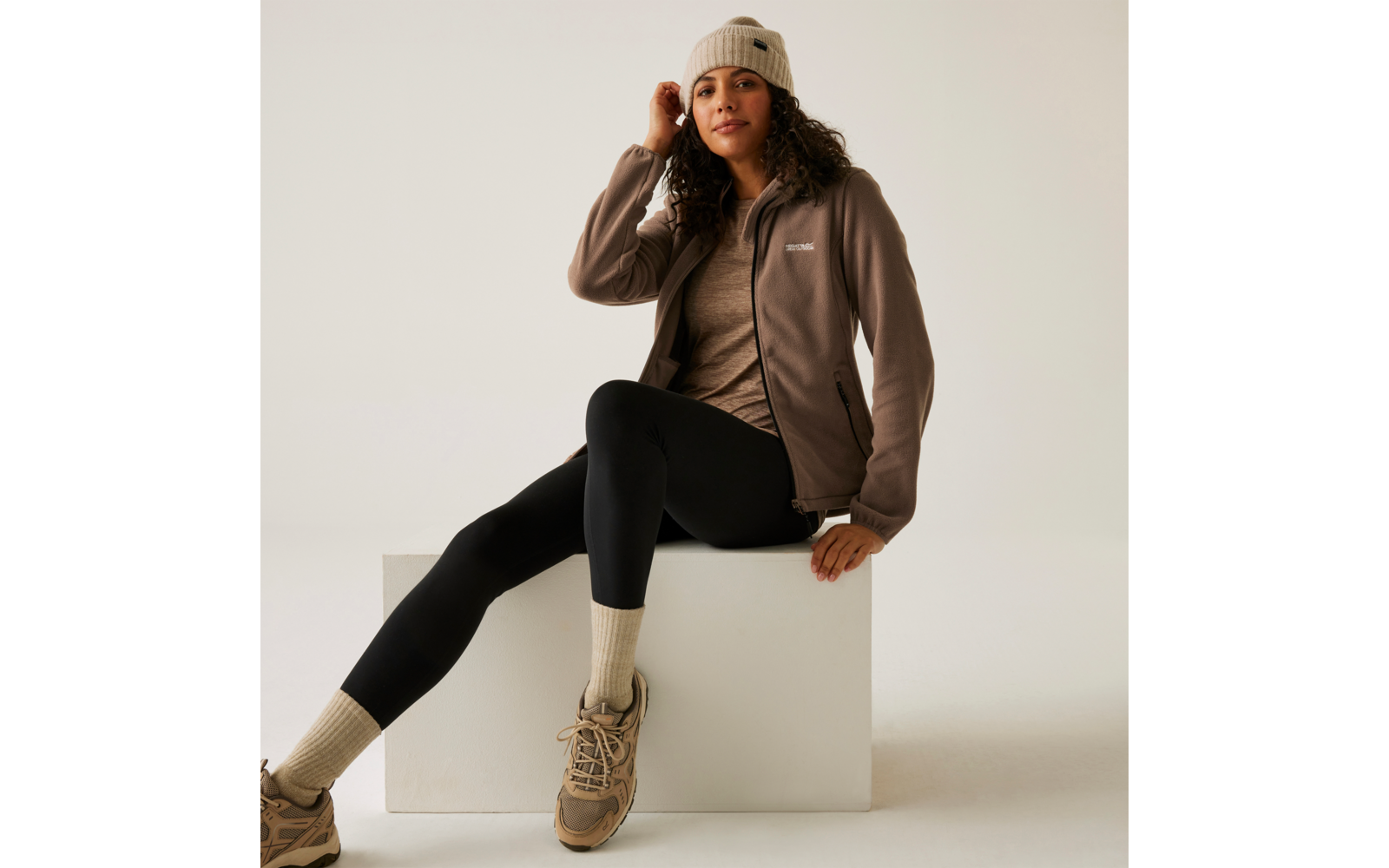 Vrouw zit op een witte kubus, draagt een bruine jas, zwarte legging en beige laarzen, lichtbeige muts