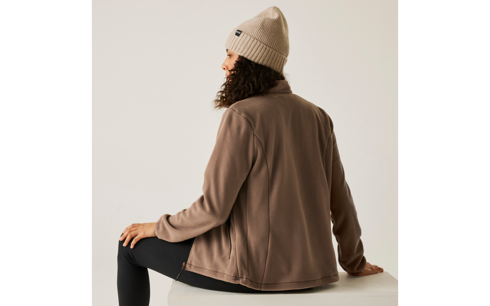 Vrouw met beige muts en bruin jack zittend zijwaarts op een witte stoel tegen een lichte achtergrond