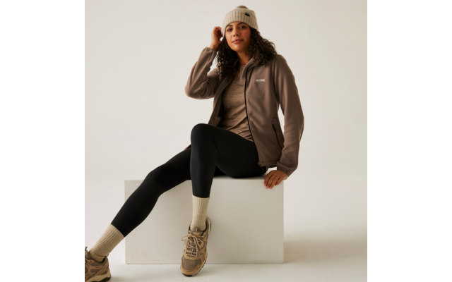 Vrouw zit op een witte kubus, draagt een bruine jas, zwarte legging en beige laarzen, lichtbeige muts