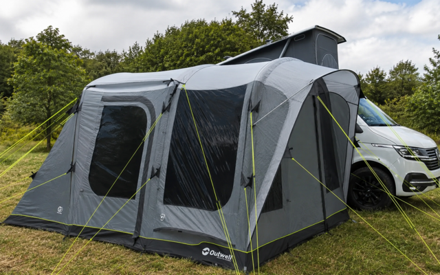 Tenda grigia fissata al lato di un camper bianco su un prato, grandi finestre scure, tiranti gialli, alberi sullo sfondo, cielo nuvoloso