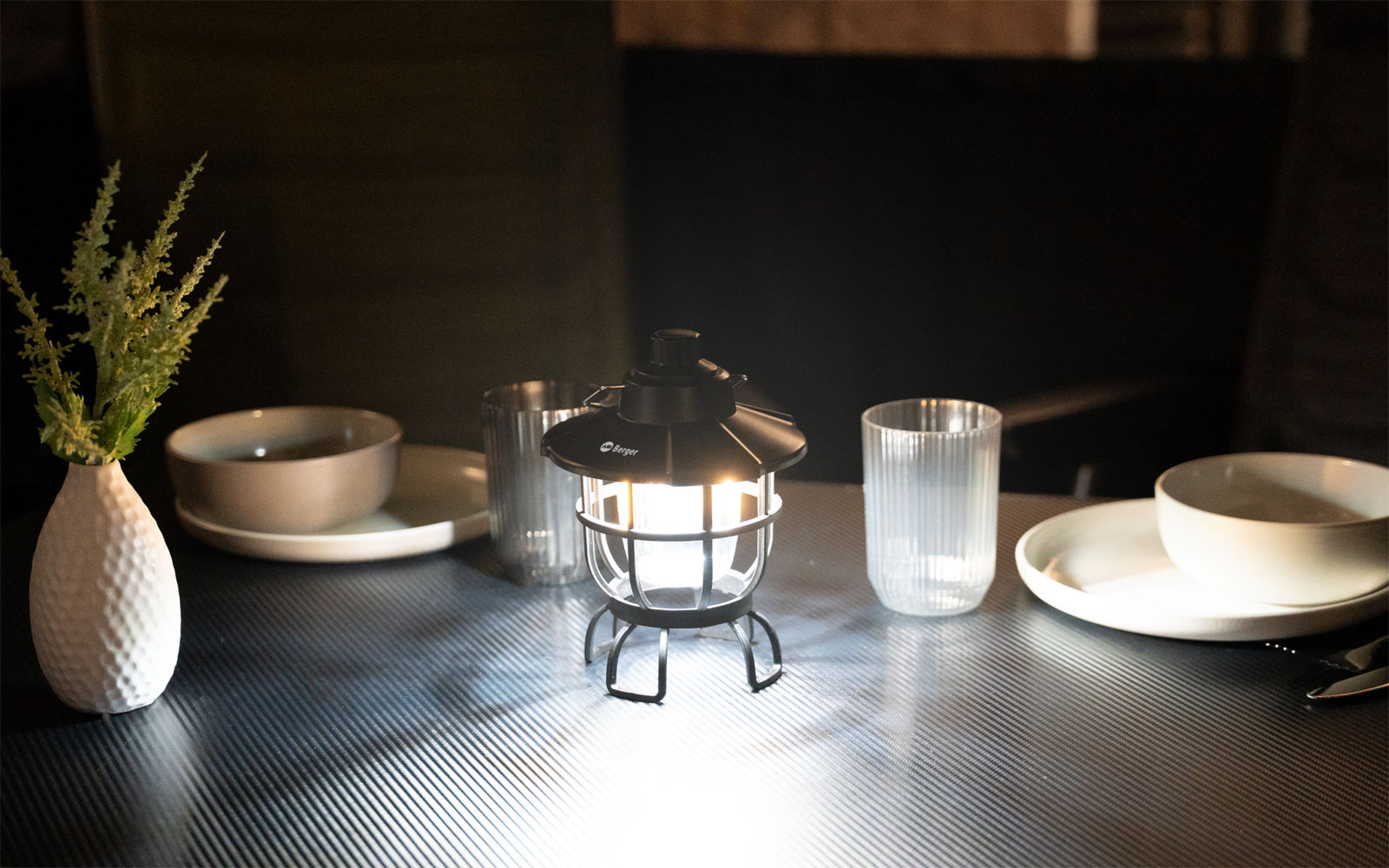 Gitterförmige Laterne mit eingeschaltetem Licht auf einem Tisch; links eine weiße Vase mit grünem Zweig; zwei Gedecke mit Teller, Schüssel und Glas