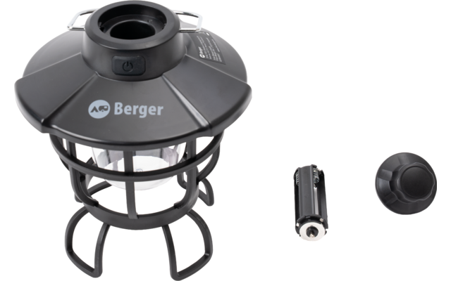 Lampe noire avec boîtier supérieur, cage métallique et lumière visible ; à côté une pile cylindrique et un cache rond en plastique
