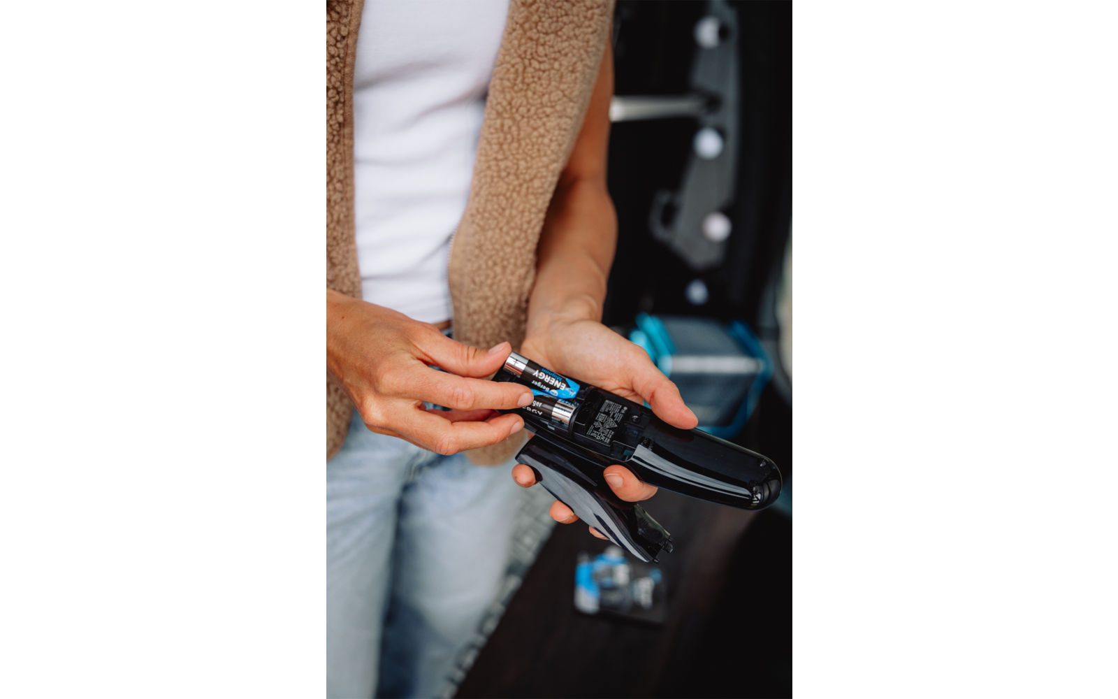 Hände halten zwei AA-Batterien an ein offenes schwarzes Batteriefach eines Geräts; Oberkörper mit weißem T-Shirt und beiger Fleeceweste, blaue Jeans, unscharfer Hintergrund mit Gegenständen