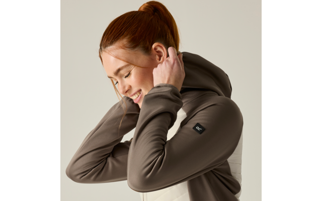 Person with red hair in a ponytail, side view, smiling, pulling up the hood of a brown hoodie; plain light background