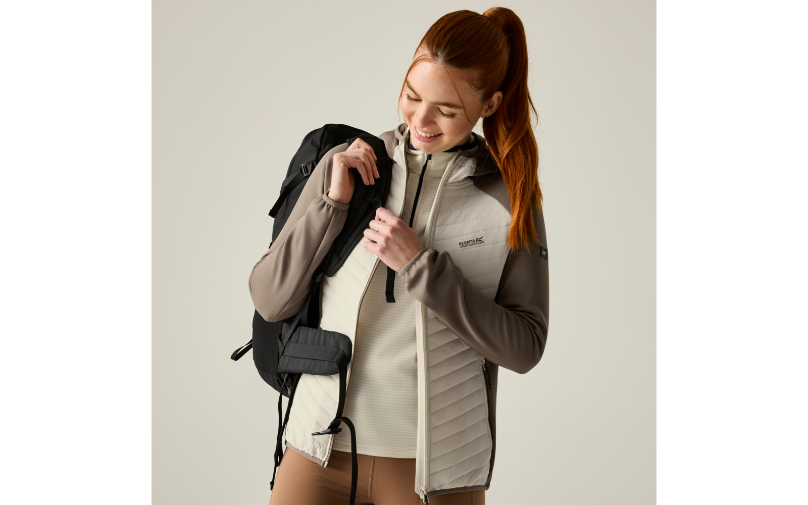Woman with red ponytail wearing a quilted outdoor jacket and backpack, holding the zipper and looking down while smiling